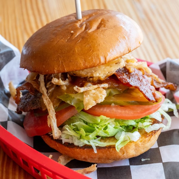 A gourmet burger with bacon, fried onions, pickles, tomato, and lettuce in a red basket.