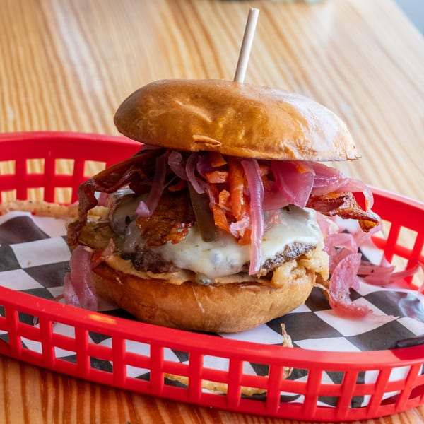 A gourmet burger with melted cheese, bacon, and pickled onions in a red basket on a wooden table.