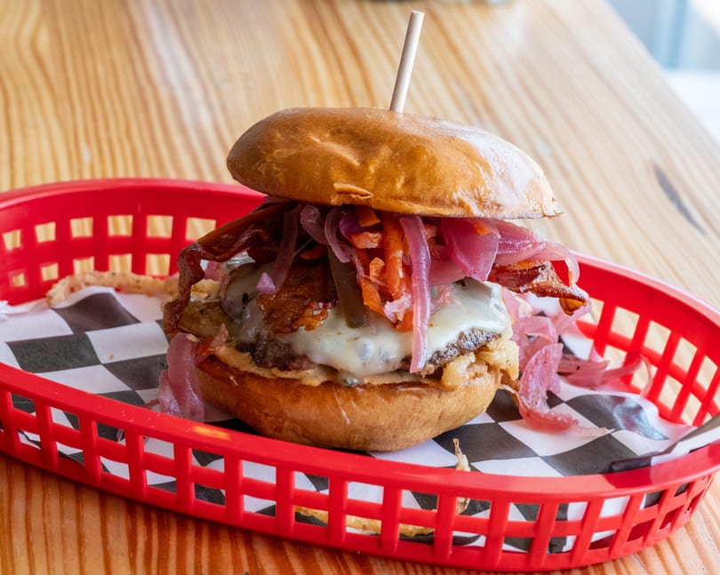 A gourmet burger with melted cheese, bacon, and pickled onions in a red basket on a wooden table.