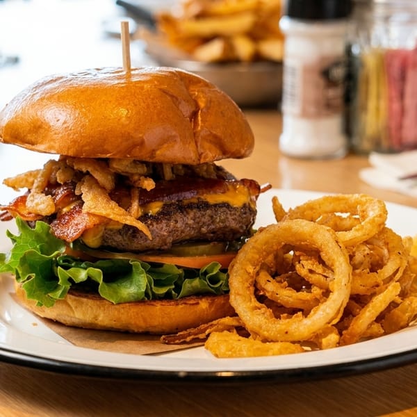 A bacon cheeseburger topped with crispy fried onions and a side of onion rings on a white plate.