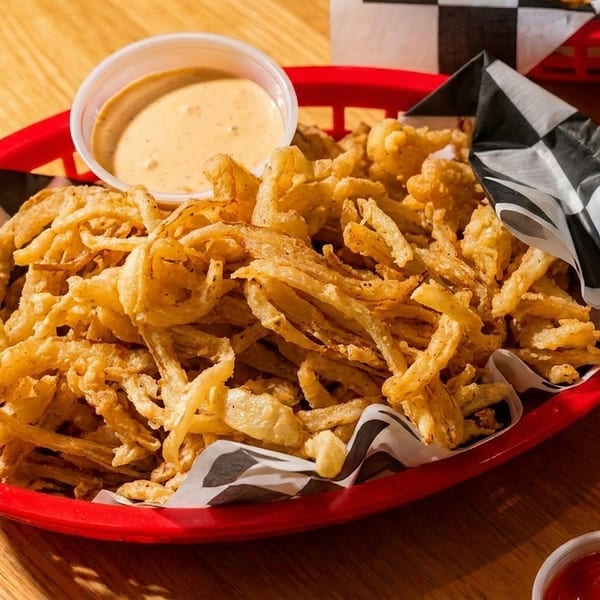 A red basket filled with crispy fried onion strings and a side of dipping sauce on a wooden table.