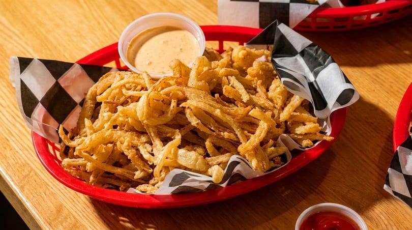 A red basket filled with crispy fried onion strings and a side of dipping sauce on a wooden table.