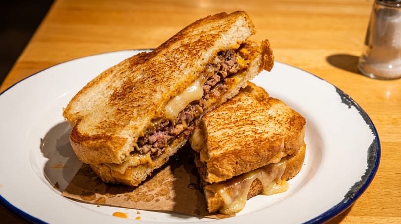 Grilled cheese sandwich with ground beef and melted cheese, cut in half on a white plate.